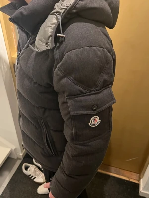 Mörkgrå pufferjacka från Moncler - Snygg mörkgrå pufferjacka från Moncler med huva och dragkedja framtill. Jackan har två sidofickor med dragkedja och en ficka med knapp på ärmen där Moncler-loggan sitter. Tjockt vadderad för extra värme och sportig look, perfekt för våren. Skann på märket fungerar men även inuti på QR koden. Hör av er för mer bilder och frågor!😊😊