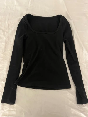 Svart långärmad topp med rundad urringning - En enkel och stilren svart långärmad topp med rundad, djup urringning. Toppen har en tight passform och är tillverkad i mjukt material som känns skönt mot huden. Perfekt att matcha med jeans eller kjol för en clean look. Väldigt mjukt mysigt material