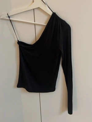 Svart oneshoulder topp Gina Tricot - Cool svart oneshoulder topp från Gina Tricot med en lång ärm och one shoulder på andra sidan (svarta tunna bandet är bara för att hänga upp). Toppens material är mjukt och stretchigt, perfekt för att skapa en snygg och unik look. Passformen är slim och toppen är enkel att matcha med olika outfits.