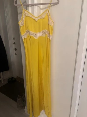 Gul långklänning med spetsdetaljer - Säljer en solgul långklänning med tunna axelband och vita spetsdetaljer vid byst och midja. Klänningen har en mjuk, följsam passform och är perfekt för sommarens alla tillfällen. Materialet känns lätt och svalt mot huden.