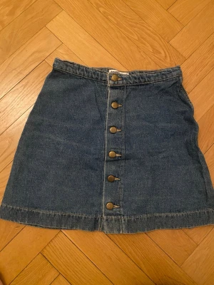 Jeanskjol med knappar American Apparel - Säljer en klassisk blå jeanskjol från American Apparel. Kjolen är kort och med snygga metallknappar framtill. Perfekt till vardags och enkel att styla med olika toppar. 