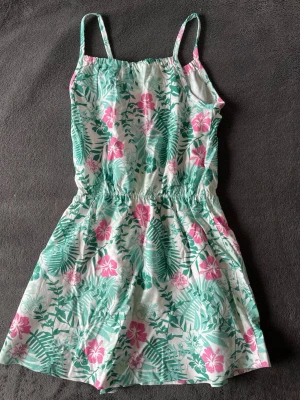 Blommig sommarklänning från Name It - Söt kortklänning från Name It med grönt och rosa blommönster på vit botten. Klänningen har smala axelband och resår i midjan för en bekväm passform. Perfekt för varma sommardagar och enkel att matcha med sandaler eller sneakers.