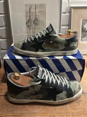 Golden goose came skor - Snygga sneakers från Golden Goose med camouflagemönster i grönt, svart och beige. Skorna har en svart stjärna på sidan, vita slitna skosnören och vit sula. Märket syns tydligt på plösen och sidan. Tillverkade i canvas och mocka för en cool streetstil.