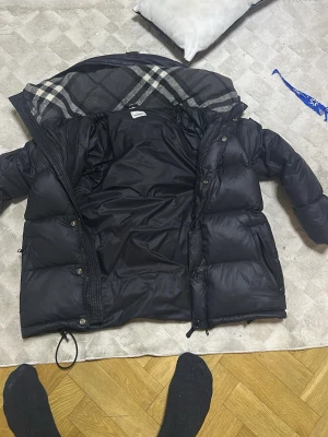 Svart dunjacka med rutigt foder - Säljer en svart dunjacka med rutigt foder i huvan. Jackan har dragkedja och knappar framtill, två sidofickor med dragkedja och en justerbar nederkant. Perfekt för kalla vinterdagar och riktigt varm. Klassisk design som passar till många stilar använd bara under den vinter helt ny den är liten på mig , pris kan diskuteras så skriv 😊😊😊.