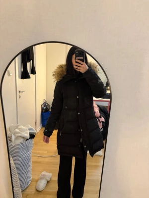 Svart parkas med fuskpäls - Svart lång parkas med huva och fuskpäls runt kragen. Jackan har dragkedja och knappar framtill samt två stora fickor. Perfekt för kalla dagar och har en klassisk, stilren look. Hollies jacka vinter 