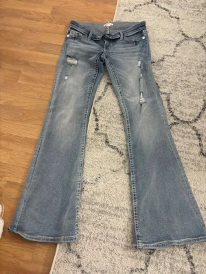Ljusblå bootcut jeans med slitningar - Säljer ett par ljusblå bootcut jeans med slitningar och hål på framsidan. Jeansen har broderade detaljer på bakfickorna och klassisk femficksmodell. Perfekta för dig som gillar en avslappnad och trendig look. 
