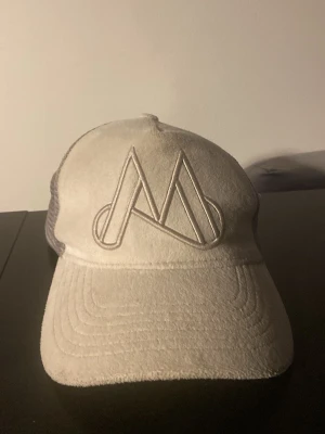 Beige truckerkeps med M-logga - Säljer en beige truckerkeps med stor broderad M-logga framtill. Kepsen har böjd skärm och mesh på sidorna och bak för extra ventilation. Perfekt för dig som gillar stilrena accessoarer med streetkänsla. Kan diskutera pris.