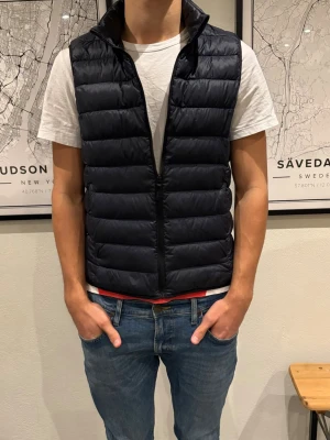 Uniqlo dunväst - Snygg svart dunväst med horisontella sömmar och hög krage. Västen har dragkedja framtill och en stilren, minimalistisk look som passar perfekt över en t-shirt eller hoodie. Materialet är lätt och ger en modern siluett. Sitter lite mer som en XS än S. Jag är 178 cm och väger 65 kg.