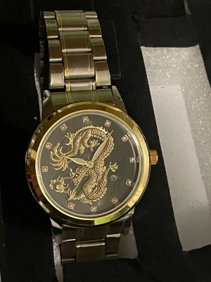 Guldig klocka med drakmotiv - Stilren klocka i guld- och silverfärgad metall med ett detaljerat drakmotiv på urtavlan. Klockan har länkarmband och markerade timmarkeringar med små stenar. Perfekt accessoar för dig som vill sticka ut.