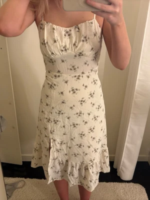 Blommig vit klänning med volang - Söt vit klänning med tunna axelband och blommigt mönster. Klänningen har en volang längst ner och en liten slits framtill. Perfekt för varma dagar och har en feminin, figurnära passform.