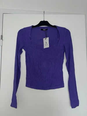 Lila ribbad topp från Bik bok - Trendig lila topp från Bik bok storlek S med ribbad struktur och fyrkantig urringning. Långärmad och figurnära passform som ger en snygg siluett. Perfekt att styla med jeans eller kjol för en cool look.