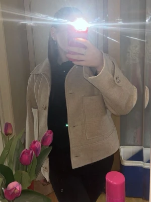 Beige ullkappa med stora knappar - Snygg beige kappa i ullmix med stora dekorativa knappar och bred krage. Modellen är kortare och har två stora fickor framtill. Perfekt för dig som vill ha en stilren och trendig look till vardags.