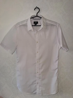 Vit kortärmad skjorta från H&M - Klassisk slimfit vit skjorta från H&M med korta ärmar och knappar framtill. Perfekt för en clean och fräsch look.