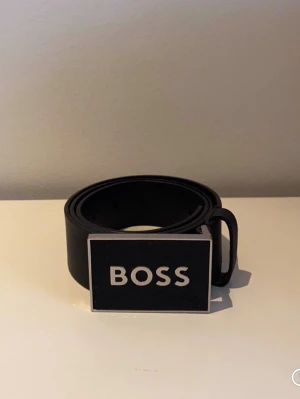 Svart skärp från BOSS med metallspänne - Stilrent svart skärp från BOSS med ett stort metallspänne där loggan är tydligt ingraverad. Skärpet har en klassisk design och är tillverkat i skinn, perfekt för att lyfta din outfit med en exklusiv touch.