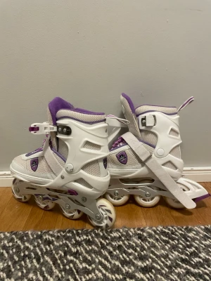 Justerbara inlines 33-37 - Säljer ett par justerbara inlines i vitt och lila med plastskal. Skorna har spännen för enkel påtagning och är designade för att passa storlek 33-37. Perfekta för dig som vill rulla fram med stil :)