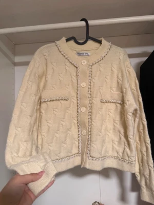 Elegant stickad kofta med pärldetaljer – klassisk & chic - Superfin krämfärgad stickad kofta från Chance Off med vackra pärldetaljer längs knäppningen, halsringningen och fickorna. En riktigt feminin och lyxig modell som ger en tidlös, CC inspirerad känsla.   Perfekt att bära: • över en klänning • till jeans för en mer uppklädd vardagslook • till kjol eller kostymbyxor  kofta, pärlkofta, stickad kofta, beige kofta, creme kofta, elegant kofta, vintage inspirerad, feminin stil, cardigan, preppy, old money style, klassisk kofta 