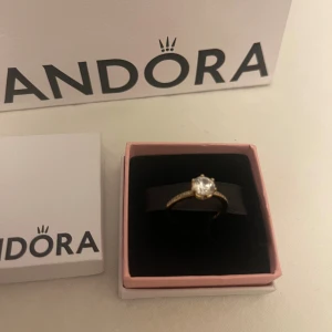 Pandora guldring - En guldfärgad ring från Pandora med en glittrande klar sten i mitten. Ringen har diamantstenar runt om (se sista bild). Kommer i original ask och påse från Pandora. Ringen är näshan helt ny, använd ett FÅÅ tal gånger! Storlek 52 med diameter 16,5mm. 