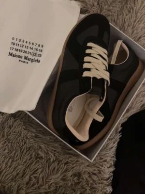 Maison Margiela svarta sneakers - Maison Margiela sneakers i svart med beige detaljer och vita skosnören. Klassisk låg modell med gummisula och stilren design. Materialmix av läder och mocka ger en lyxig känsla. Perfekta för dig som gillar minimalistisk och trendig stil.