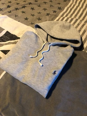 Grå hoodie från Polo Ralph Lauren - Säljer en klassisk grå hoodie från Polo Ralph Lauren med vit dragsko i huvan och den ikoniska lilla blå loggan broderad på bröstet. Hoodien har känguruficka framtill och är gjord i mjukt bomullsmaterial. Perfekt för en chill och stilren look.