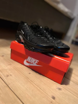 Nike Air Max Plus svarta sneakers - Säljer ett par svarta Nike Air Max Plus sneakers med mesh- och syntetmaterial. Skorna har den ikoniska Tuned Air-sulan och vita detaljer på sidan. Perfekta för dig som gillar streetwear och vill ha en sportig look.