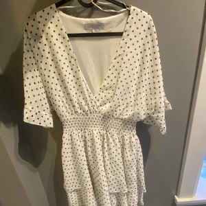 Vit prickig omlottklänning - Söt vit omlottklänning med svarta prickar och smock i midjan. Klänningen har korta ärmar med lätt volang och v-ringad hals. Perfekt för dig som gillar en feminin och trendig stil.
