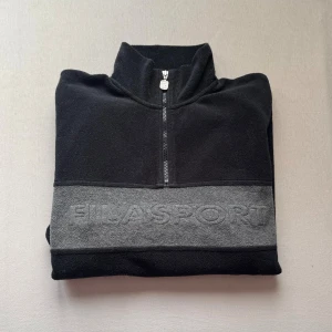 Svart fleece från FILA SPORT - Svart stickad fleece med halv dragkedja från FILA SPORT. Tröjan har en bred grå panel över bröstet med broderad logga, hög krage och lång ärm. Perfekt för lager på lager och chill dagar. Materialet är mjukt och värmande.