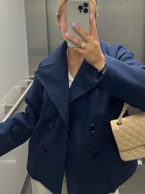 Mörkblå skepparkavaj med bred krage - Stilren mörkblå skepparkavaj med dubbelknäppning och bred krage. Jackan har en klassisk, boxig passform och stora svarta knappar framtill. Perfekt för dig som gillar en tidlös look. Materialet ser ut att vara ullblandning.