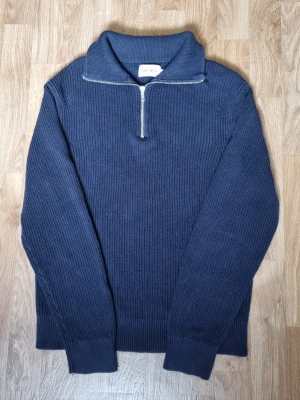 Mörkblå stickad tröja med halvzip - Snygg mörkblå stickad tröja från East West med halvzip och hög krage. Tröjan har ribbade muddar och en diskret struktur i stickningen. Perfekt för lager på lager och enkel att matcha med jeans eller chinos.