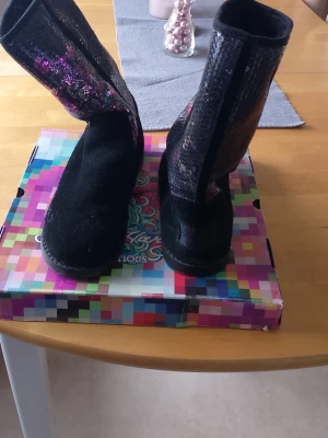 Svarta boots med paljetter Ed Hardy märke.  - Säljer ett par svarta boots med glittriga paljetter på skaftet. Skorna har rund tå och mjuk sula, perfekta för dig som vill sticka ut lite extra. Materialet är en mix av textil och syntet med en skimrande finish på ovandelen.