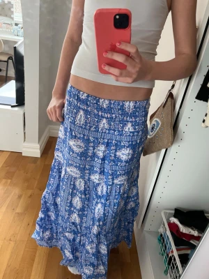 Blå långkjol från Gina Tricot - Säljer en blå långkjol från Gina Tricot med vitt blommigt mönster. Kjolen har smock i midjan för skön passform och är gjord i ett lätt och luftigt material, perfekt för varma dagar. Flowy modell som ger en avslappnad och trendig look.