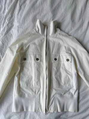 Vit jacka från Gant - Snygg vit bomberjacka från Gant med dragkedja framtill och två stora bröstfickor med lock och knappar. Jackan har ribbade muddar vid krage och nederkant samt broderad text på insidan. Perfekt för dig som gillar stilren och klassisk design. Säljer min jätte fina Gant jacka som inte kommer till nån större användning💞💞💞 finns en fläck som bild 4 visar Är en M men skulle säga att de passar en S/XS🩷