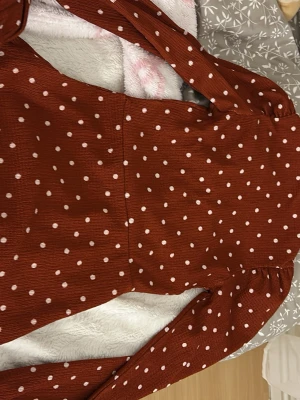 Rostbrun prickig omlottopp - Supersöt rostbrun omlottopp med vita prickar och lång ärm. Toppen har v-ringning och markerad midja som ger en snygg siluett. Perfekt att styla med jeans eller kjol för en trendig look.