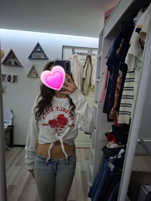 Vit croppad långärmad topp - Säljer en vit croppad långärmad topp med röd text och rosor framtill. Toppen är dock klippt framtill så man kan knyta och men väldigt fin och skön att ha på sig. Perfekt för dig som gillar trendiga och bekväma plagg.