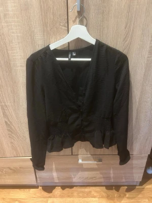 Svart v-ringad blus med volang - Trendig svart blus med v-ringning och långa ärmar. Blusen har smockdetaljer i midjan och volang nedtill för en snygg siluett. Tillverkad i ett lätt, något glansigt tyg som ger en elegant känsla. Perfekt att styla med jeans eller kjol.