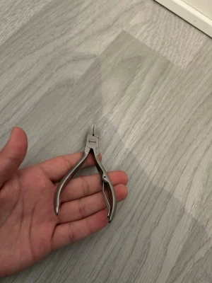Nagelklippare i rostfritt stål - Nagelklippare i silverfärgat rostfritt stål med ergonomiskt handtag och vass, böjd klipptopp. Perfekt för att trimma naglar och nagelband med precision. Enkel att använda och lätt att rengöra.