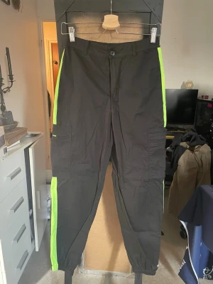 Svarta cargobyxor med neonränder - Säljer ett par svarta cargobyxor från Forever 21 med neongröna ränder längs sidorna. Byxorna har flera fickor, resår vid benslut och en avslappnad passform. Perfekta för en streetwear-look och riktigt bekväma att bära.