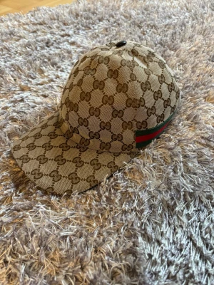 Gucci beige keps med GG-mönster - Säljer en beige keps från Gucci med det klassiska GG-monogrammet över hela kepsen🤝 Kepsen har en böjd skärm, justerbart band bak i brunt läder och en grön-röd rand på sidan. Materialet är canvas och detaljerna ger en lyxig känsla. Mycket bra skick och självklart äkta✅