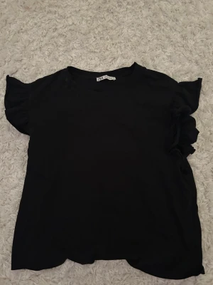Svart topp med volangärmar från Zara - Säljer en svart topp från Zara med korta volangärmar. Toppen har en enkel, rund halsringning och är gjord i mjukt bomullstyg. Perfekt att styla med jeans eller kjol för en clean look.