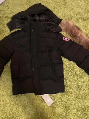 Svart dunjacka från Canada Goose - Säljer en svart dunjacka från Canada Goose med avtagbar huva och pälskant. Jackan har två stora framfickor, dragkedja och knappar framtill samt den klassiska logotypen på ärmen. Perfekt för kalla vinterdagar.