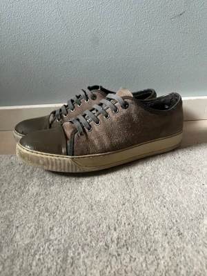 Lanvin skor  - Snygga bruna sneakers med ovandel i mocka och tå i blankt gummi. Skorna har grå skosnören och en beige sula som ger en retrovibe. Perfekta för dig som gillar en stilren men ändå unik look. Alla skador är synliga på bilderna och är inte så farliga. 