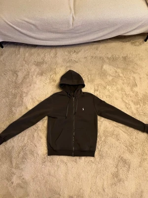Svart hoodie från Polo Ralph Lauren - Säljer en svart hoodie från Polo Ralph Lauren med dragkedja framtill och klassisk huva med snören. Tröjan har två fickor och den ikoniska vita Polo-loggan broderad på bröstet. Perfekt för en chill och stilren look.