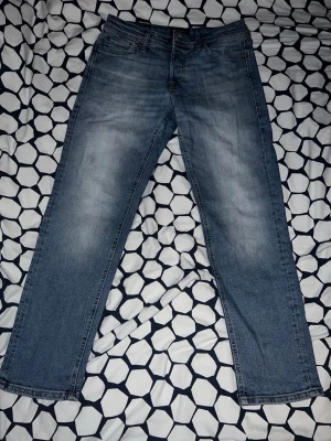 Blå raka jeans med slitningar - Säljer ett par klassiska blå jeans med raka ben och snygga slitningar på framsidan. Jeansen har fem fickor och en läderpatch bak i midjan. Perfekta till en avslappnad stil och passar till det mesta. Materialet är denim i bomull. Storleken är W=30 L=30