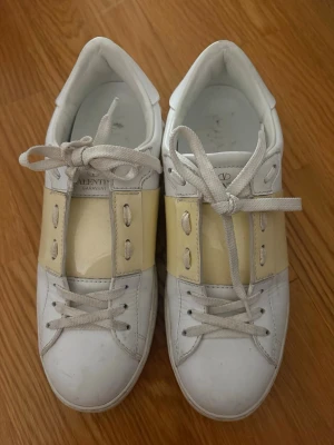Vita sneakers från Valentino Garavani - Snygga vita sneakers från Valentino Garavani med bred ljusgul rem över mitten och vita skosnören. Skorna har en klassisk låg siluett och är tillverkade i läder med detaljer i gummi. Perfekta för dig som gillar stilrena och exklusiva sneakers. De är köpta second hand i Paris därav kan jag inte garantera att de är äkta