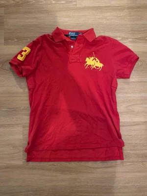 Röd pikétröja från Polo Ralph Lauren - Klassisk röd pikétröja från Polo Ralph Lauren med gul broderad logga på bröstet och siffran 3 på ärmen. Tröjan har krage, knappar framtill och är tillverkad i bomull. Custom fit-modell som ger en snygg passform.