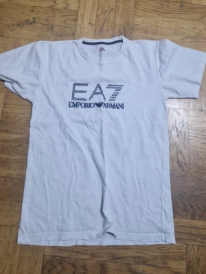 Vit EA7 Emporio Armani t-shirt - Snygg vit t-shirt från EA7 Emporio Armani med tryck på bröstet. Klassisk passform och rund halsringning. Perfekt till jeans eller shorts för en clean och stilren look.