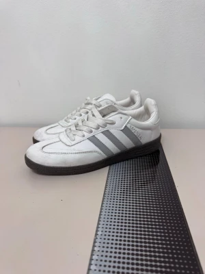 Vita Adidas Samba sneakers - Klassiska vita Adidas Samba sneakers med gråa ränder på sidorna och mörk sula.behöver endast lite rengöring annars är dom nästen till nya  Skorna har lågt skaft, snörning och är tillverkade i läder och mocka. Perfekta för dig som gillar retrostil och sportig vibe. Obs original box följer inte med 