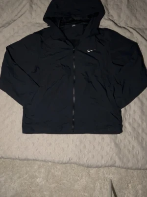 Svart vindjacka med huva från Nike - Snygg svart vindjacka från Nike med dragkedja framtill och huva. Jackan har en minimalistisk design med Nike-logga på bröstet och är tillverkad i ett lätt, vindtätt material. Perfekt för blåsiga dagar och enkel att matcha med olika outfits.