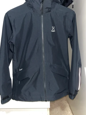 Haglöfs vindjacka - Snygg svart regnjacka från Haglöfs med huva och hel dragkedja framtill. Jackan har två stora fickor med lock, reflexdetaljer på ärmarna och justerbara muddar. Tillverkad i slitstarkt, vattenavvisande material som passar perfekt för regniga och vindiga dagar. Köpt 1200kr pris kan diskuteras!