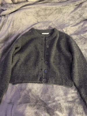 Mörkgrå cropped kofta med knappar - Mysig mörkgrå cropped kofta med rund halsringning och stora knappar framtill. Koftan har långa ärmar och bred ribbstickad kant nedtill. Perfekt att slänga över en t-shirt eller klänning för en chill look.