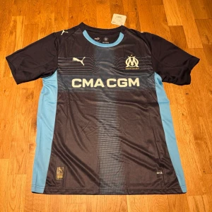 Marseille away kit 25/26 - Snygg svart och ljusblå matchtröja från Olympique de Marseille med Puma-logga och klubbmärke på bröstet. Tröjan har korta ärmar, rund hals och tryckt sponsorlogga framtill. Tillverkad i lätt och ventilerande funktionsmaterial.